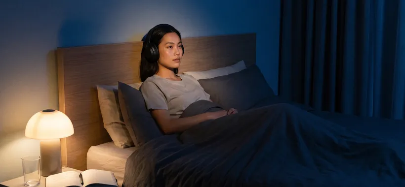 A person sits awake in bed at night with headphones on, auditioning a spoken recording by lamplight, a dream journal open on the nightstand.
