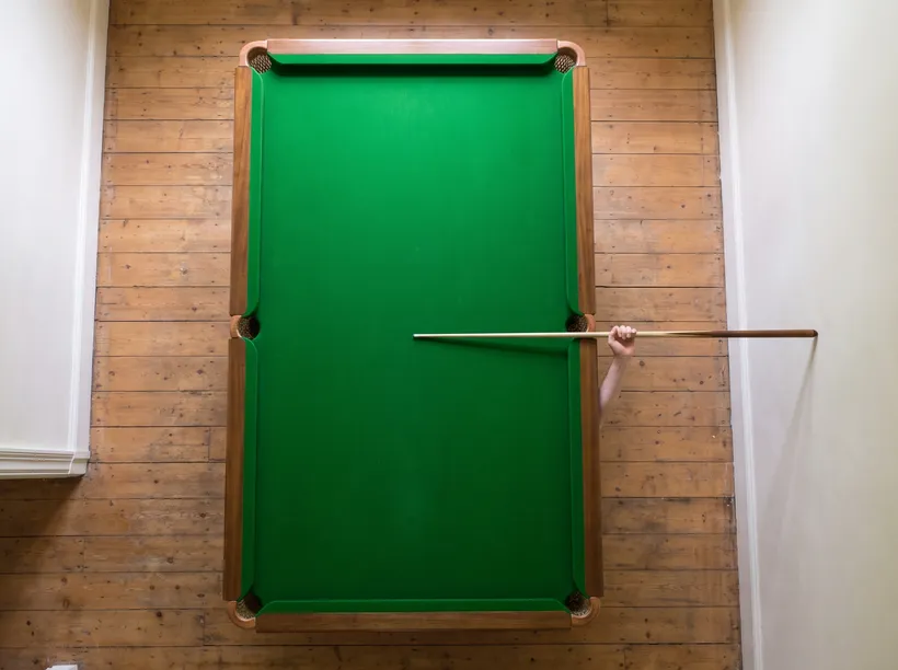 Overhead view of a snooker table in a room with one full-length cue swung straight out from the table edge, its tip reaching the adjacent wall